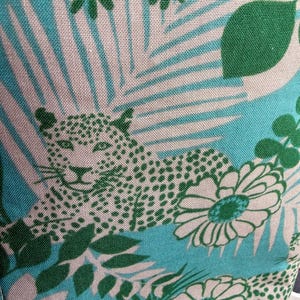May include: A fabric textile featuring a repeating pattern of green leopards, white daisies, and green leaves set against a bright turquoise background. The design includes stylised palm fronds and floral elements printed in a crisp, clear graphic style.