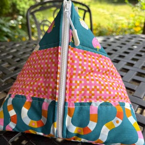 May include: A pyramid-shaped fabric pouch featuring a white zip closure. The exterior displays a teal section with pink strawberry motifs, a central band with a pink and orange grid pattern, and a bottom section with a teal, orange, and white wavy design.