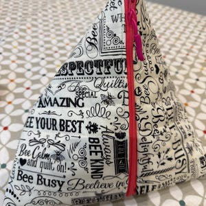 May include: A triangular fabric pouch with a white background and black text, including phrases like "Bee Calm" and "Bee Busy." The pouch has a bright pink zip and is likely used for storage or as a small accessory. The fabric is patterned with bees and quilting-related words.
