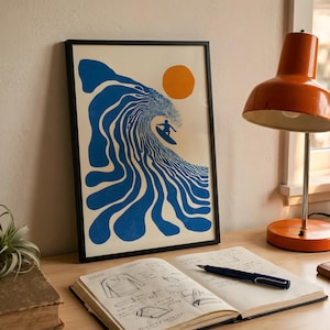 May include: Framed art print featuring a surfer riding a large blue wave under an orange sun. The artwork has a minimalist, retro style. The print is in a black frame and is displayed on a wooden desk.