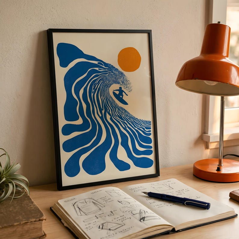 May include: Framed art print featuring a surfer riding a large blue wave under an orange sun. The artwork has a minimalist, retro style. The print is in a black frame and is displayed on a wooden desk.