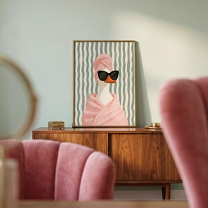 May include: A framed wall art print featuring a white duck wearing a pink towel wrapped around its head and body, paired with black cat-eye sunglasses. The background consists of vertical wavy lines in shades of light green and white.