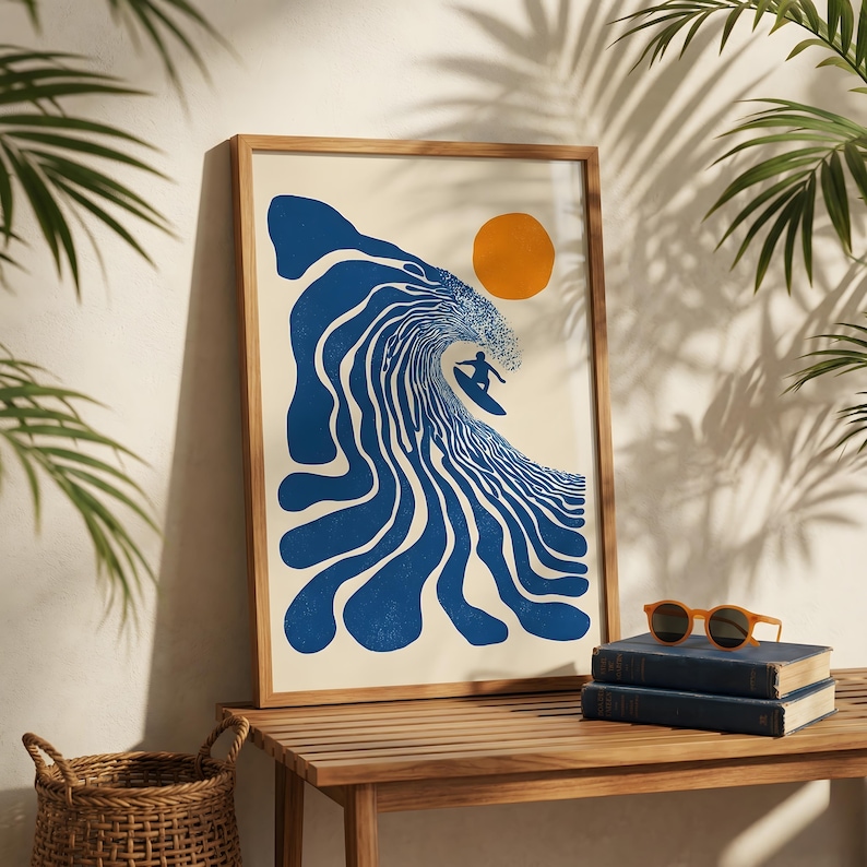 May include: A framed art print featuring a stylized blue wave with a surfer and an orange sun. The artwork is in a wooden frame and rests on a wooden table. Palm fronds and a woven basket are also visible.