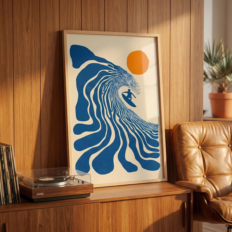 May include: Framed art print featuring a surfer riding a large blue wave under an orange sun. The artwork has a retro, minimalist style, perfect for coastal decor. The print is in a wooden frame.