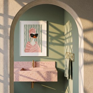 May include: A framed wall art print featuring a white duck wearing black sunglasses and a pink towel wrapped around its head and body, set against a background of vertical teal and white wavy stripes, displayed in a bathroom setting.