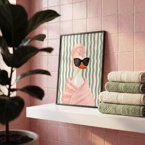 May include: A framed wall art print featuring a white goose wearing black cat-eye sunglasses and a pink towel wrapped around its head and body, set against a background of vertical wavy teal and white stripes in a clean, modern composition.