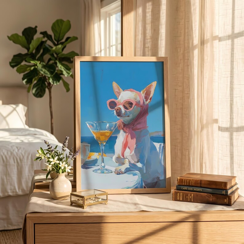 Puede incluir: Una impresi&oacute;n art&iacute;stica enmarcada representa a un chihuahua con gafas de sol rosas y una bufanda, disfrutando de un c&oacute;ctel. El perro est&aacute; sentado en una mesa con una copa de martini, sobre un fondo de cielo azul y oc&eacute;ano. La obra de arte se exhibe sobre una mesa de madera con una peque&ntilde;a caja de joyas y un jarr&oacute;n de flores.