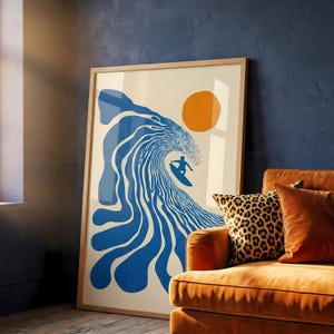 May include: A framed art print featuring a surfer riding a blue wave under an orange sun. The artwork has a minimalist style with a cream background and is displayed in a room with an orange couch and leopard print pillow.