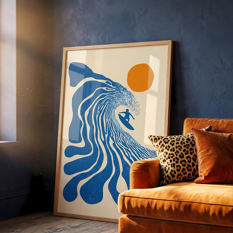 May include: A framed art print featuring a surfer riding a blue wave under an orange sun. The artwork has a minimalist style with a cream background and is displayed in a room with an orange couch and leopard print pillow.