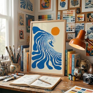 May include: A framed art print featuring a surfer riding a blue wave under an orange sun. The artwork is displayed on a wooden desk with art supplies, books, and a vintage camera. The print is surrounded by other art pieces.