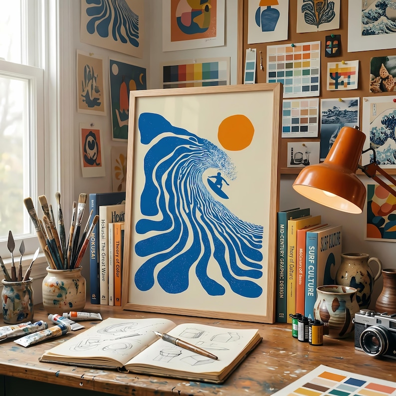 May include: A framed art print featuring a surfer riding a blue wave under an orange sun. The artwork is displayed on a wooden desk with art supplies, books, and a vintage camera. The print is surrounded by other art pieces.