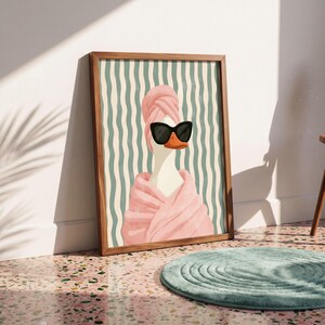May include: A framed wall art print featuring a white goose wearing black sunglasses and a pink towel wrapped around its head and body. The background consists of vertical wavy lines in alternating shades of light green and cream.