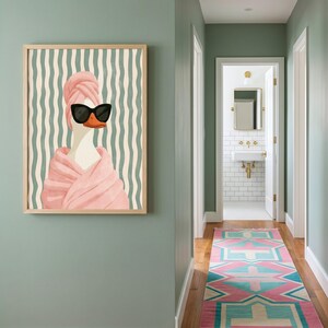 May include: A framed wall art print featuring a white duck wearing black sunglasses and a pink towel wrapped around its head and body. The background consists of vertical wavy lines in shades of light green and cream, set in a wooden frame.