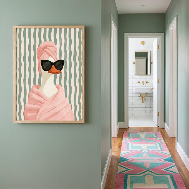 May include: A framed wall art print featuring a white duck wearing black sunglasses and a pink towel wrapped around its head and body. The background consists of vertical wavy lines in shades of light green and cream, set in a wooden frame.