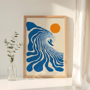 May include: A framed art print featuring a surfer riding a large blue wave under an orange sun. The artwork has a minimalist style with a cream-colored background and a wooden frame. The surfer is a silhouette.