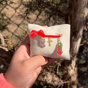 May include: A small, cream-colored pillow with a red bow and a gold safety pin holding three charms. The charms include a foot with a blue eye, a gold cross, and a colorful depiction of the Virgin Mary.