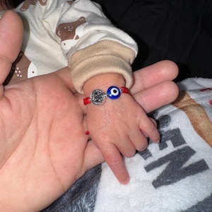 Peut inclure: Un bracelet en fil rouge avec un charm argenté et une perle en forme d'œil bleu est porté au poignet d'un bébé. La main du bébé est tenue par une main d'adulte. Le bébé porte un body blanc et beige.