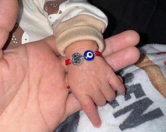 May include: A red string bracelet with a silver charm and a blue eye bead is worn on a baby's wrist. The baby's hand is held by an adult hand. The baby is wearing a white and tan onesie.