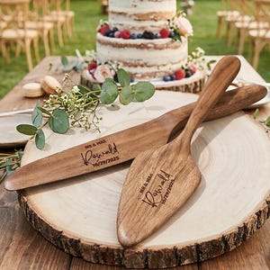 Puede incluir: Un juego de cuchillo y servidor de pastel de madera con la inscripción "MR & MRS, Ranwald, 10/29/2025" sobre una rodaja de madera. Un pastel de dos niveles con bayas y flores se encuentra en el fondo, en un entorno de ceremonia de boda.