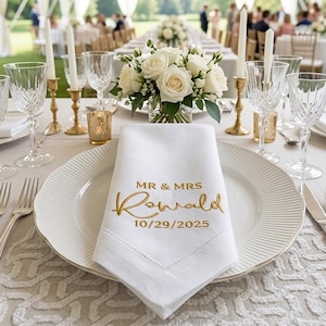 May include: A white cloth napkin featuring gold embroidered text that reads MR & MRS Rowald and the date 10/29/2025. The napkin is folded on a white ribbed dinner plate, surrounded by crystal glassware, gold candlesticks, and a floral centrepiece.