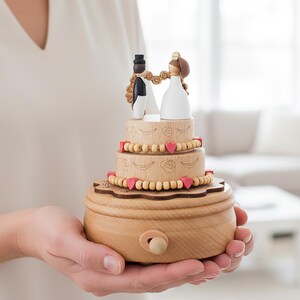以下が含まれることがあります： 2体の小さなフィギュアが乗った段重ねのケーキデザインの木製オルゴール。ケーキは明るい木製のビーズ、小さなピンクのハートのアクセント、彫刻された花柄で装飾されています。ベースの前面には小さな木製のクランクが付いています。