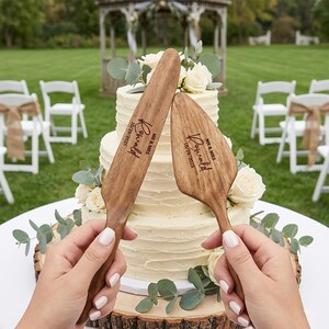 Pu&ograve; includere: Set di coltello e paletta per torta in legno con testo inciso. Il coltello e la paletta sono tenuti davanti a una torta bianca a pi&ugrave; strati decorata con rose bianche e verde. Il testo sul coltello e sulla paletta recita "Ronald" e "10/29/2025".