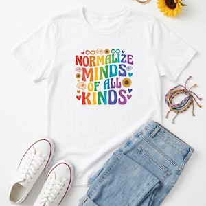 Puede incluir: Una camiseta blanca de manga corta con un estampado gráfico colorido que dice NORMALIZE MINDS OF ALL KINDS en una fuente ondulada, rodeado de pequeños iconos de cerebros, girasoles, símbolos de infinito y corazones de colores.