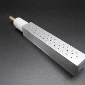 Op de afbeelding: Een rechthoekig, zilverkleurig metalen object met gelijkmatig verdeelde gaten. Het heeft een witte cilindrische component aan één uiteinde en een goudkleurige metalen staaf die zich ervan uitstrekt. Het object ligt op een donkere ondergrond.