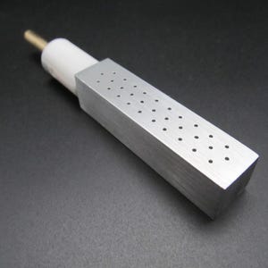 Può includere: Uno strumento metallico argentato con un corpo rettangolare perforato con piccoli fori. Ha una base cilindrica bianca e un'asta color oro. Lo strumento è progettato per uno scopo specifico.