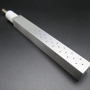 Può includere: Un oggetto metallico rettangolare color argento con fori distanziati uniformemente sulla superficie superiore. Ha una sezione in ceramica bianca e una punta dorata. L'oggetto è posto su uno sfondo scuro.