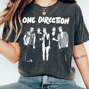 Może przedstawiać: Ciemnoszara koszulka z białym napisem "ONE DIRECTION" nad czarno-białym zdjęciem członków zespołu. Koszulka ma wygląd vintage, z efektem zużycia.