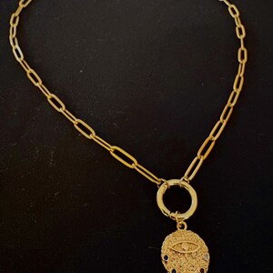 May include: Gold-toned necklace with a chain link design. The necklace features a circular pendant with an eye design and colorful gemstones. The pendant is attached to a gold ring, which is connected to the chain.