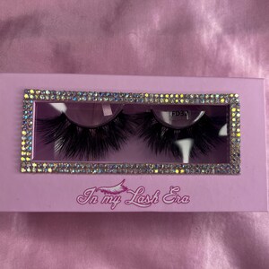 May include: A pair of black false eyelashes in a light purple box. The box has a rectangular window with a rhinestone border. The text "In my Lash Era" is printed on the box.