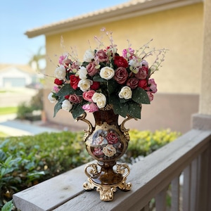 May include: A decorative vase filled with an arrangement of colourful roses. The vase is brown with gold accents and ornate handles. The flowers are red, white, pink, and burgundy, with green leaves. The vase sits on a wooden surface.