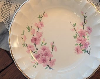 May include: A round white ceramic bowl with a scalloped edge and a gold-trimmed rim. The interior features a delicate floral pattern of pink cherry blossoms on brown branches, arranged in two distinct clusters on opposite sides.