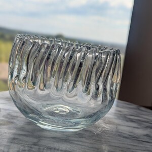 May include: A clear glass bowl featuring a distinct ribbed or fluted texture along the exterior walls. The bowl has a rounded base and a wide, open top with a scalloped edge created by the vertical glass ridges that wrap around the entire vessel.