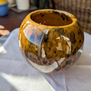 May include: A spherical ceramic vase featuring a glossy finish with a mottled pattern of deep amber, brown, and black tones. The vessel has a wide, circular opening at the top and a smooth, rounded body that reflects light across its glazed surface.