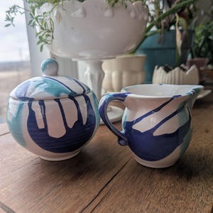 May include: A ceramic sugar bowl with a lid and a creamer. Both pieces are white with blue and teal abstract paint strokes. The sugar bowl is round, and the creamer has a handle and a spout. The set is on a wooden surface.