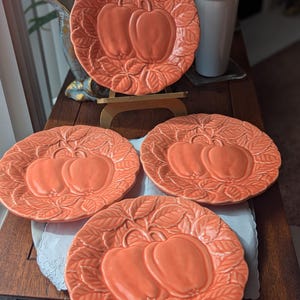 May include: Four round ceramic plates in a salmon-orange color, each featuring a raised, embossed design of two apples surrounded by detailed leaves. The plates have a scalloped edge and a glossy finish, showcasing a consistent fruit motif.