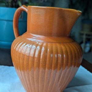 May include: An orange ceramic pitcher with a handle and a spout. The pitcher has a ribbed design on the body and a glossy finish. The pitcher is sitting on a white surface.
