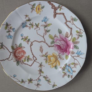 May include: White ceramic plate with a scalloped edge, featuring a floral design with pink, yellow, and blue roses, along with brown branches and green leaves. The plate has a gold rim.