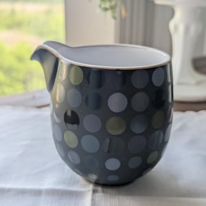 May include: A ceramic creamer pitcher featuring a dark charcoal base color adorned with a repeating pattern of circular dots in varying shades of muted blue, grey, and olive green. The vessel has a smooth, rounded body and a distinct pouring spout.
