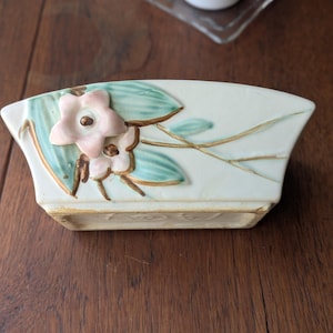 May include: A rectangular, off-white ceramic planter with a floral design. The planter features a light pink flower with brown accents and green leaves. The planter is on a dark wooden surface.