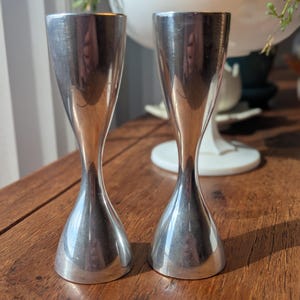 May include: Two silver-colored, hourglass-shaped shot glasses. The glasses have a flared base and top, with a narrow waist. They are displayed on a wooden surface, reflecting the light. The background includes a white decorative bowl with greenery.