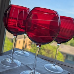 May include: Three red wine glasses with clear stems and bases. Each glass features a distinct etched pattern: one with vertical lines, one with horizontal rings, and one with fine horizontal striations. The bowls are round and deep red in color.