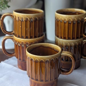 May include: A set of five brown ceramic mugs with a vertical ribbed texture and a lighter tan drip glaze along the upper rim. Each mug features a sturdy side handle and a cylindrical body, arranged in a stacked formation on a white surface.
