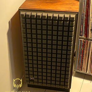 May include: A vintage speaker with a wooden top and a dark gray foam front panel. The speaker has a rectangular shape and a small JBL logo. The speaker is part of a home audio setup.