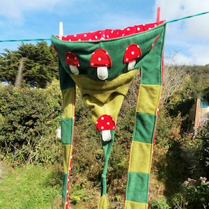 May include: A green and yellow hooded scarf with red and white mushroom appliques hanging on a clothesline. The scarf has a pointed hood and long tails.