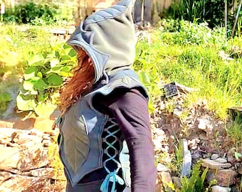 May include: A dark blue sleeveless vest featuring a pointed hood, leaf-shaped paneling on the front, and side lacing with matching blue cords. The vest is worn over a black long-sleeved shirt and paired with a long, flowing dark blue skirt.