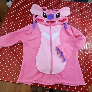 Puede incluir: Una sudadera con capucha rosa con cremallera que representa al personaje Angel de Lilo & Stitch. La sudadera tiene una capucha con orejas grandes, detalles morados y un dise&ntilde;o espec&iacute;fico del personaje en la parte delantera. Las mangas tienen un dise&ntilde;o de pu&ntilde;o &uacute;nico.